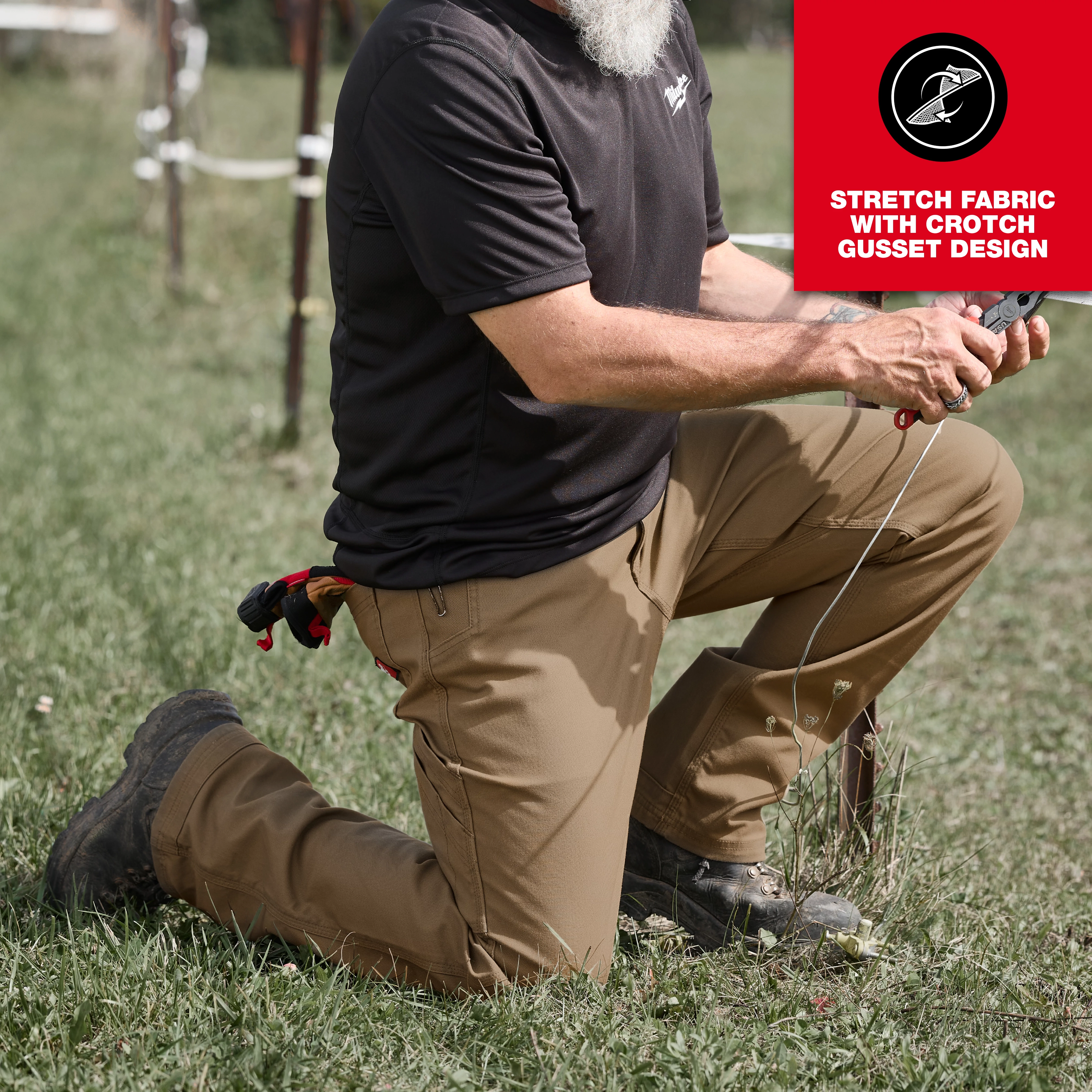 A man is kneeling outdoors, wearing tan Work Pants and a black shirt. The pants feature stretch fabric with a crotch gusset design, highlighted in a red upper right corner of the image. The person is holding a tool and has multiple items attached to their belt.