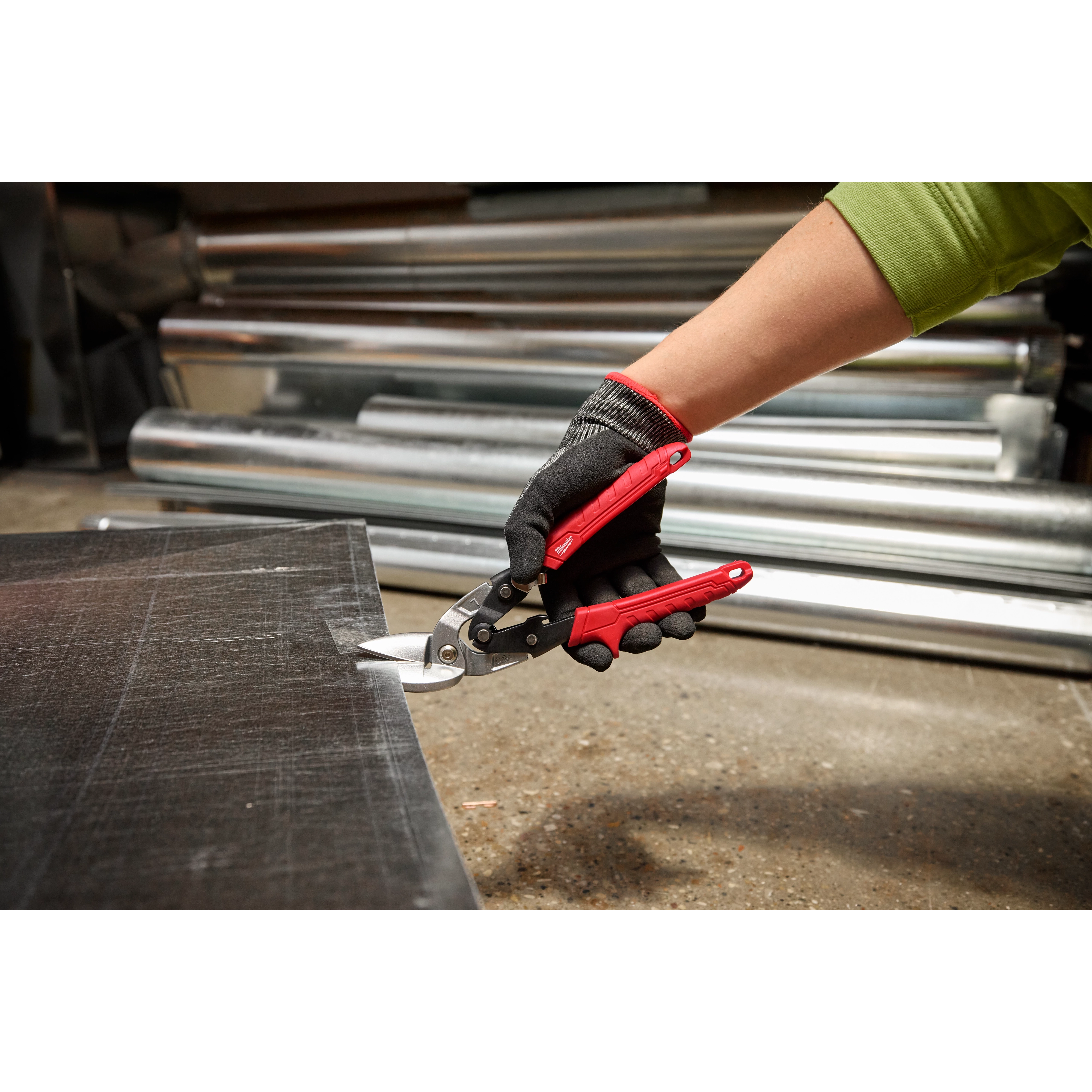 A person wearing a green sleeve and black gloves is using Left Cutting Offset Aviation Snips with red handles to cut a piece of metal sheet. Rolled metal sheets are visible in the background.