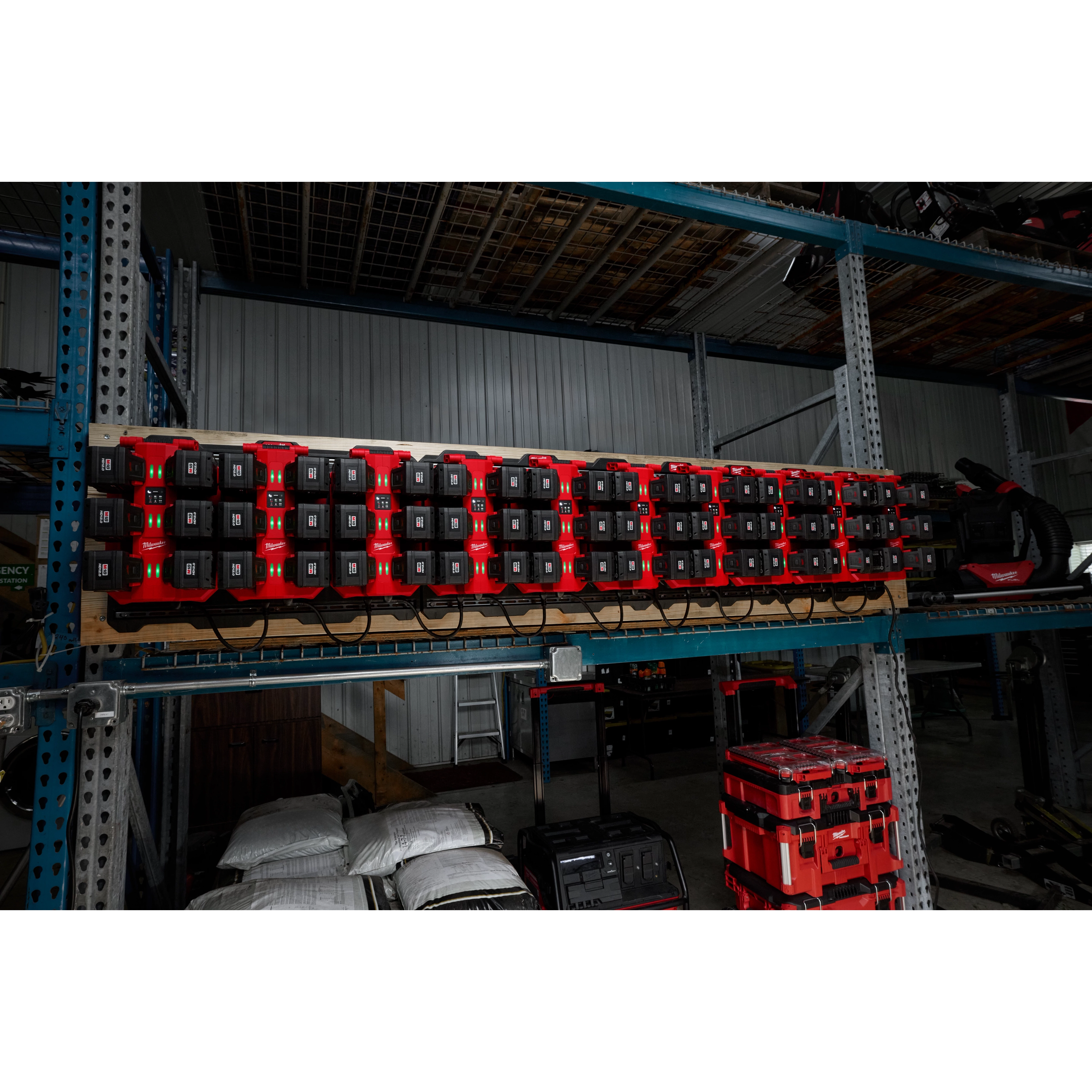 The image shows multiple M18™ Six Bay Daisy Chain Rapid Chargers mounted on a rack in a warehouse. They are charging numerous battery packs simultaneously. The chargers are red and black, and the shelf below holds various tools and equipment.