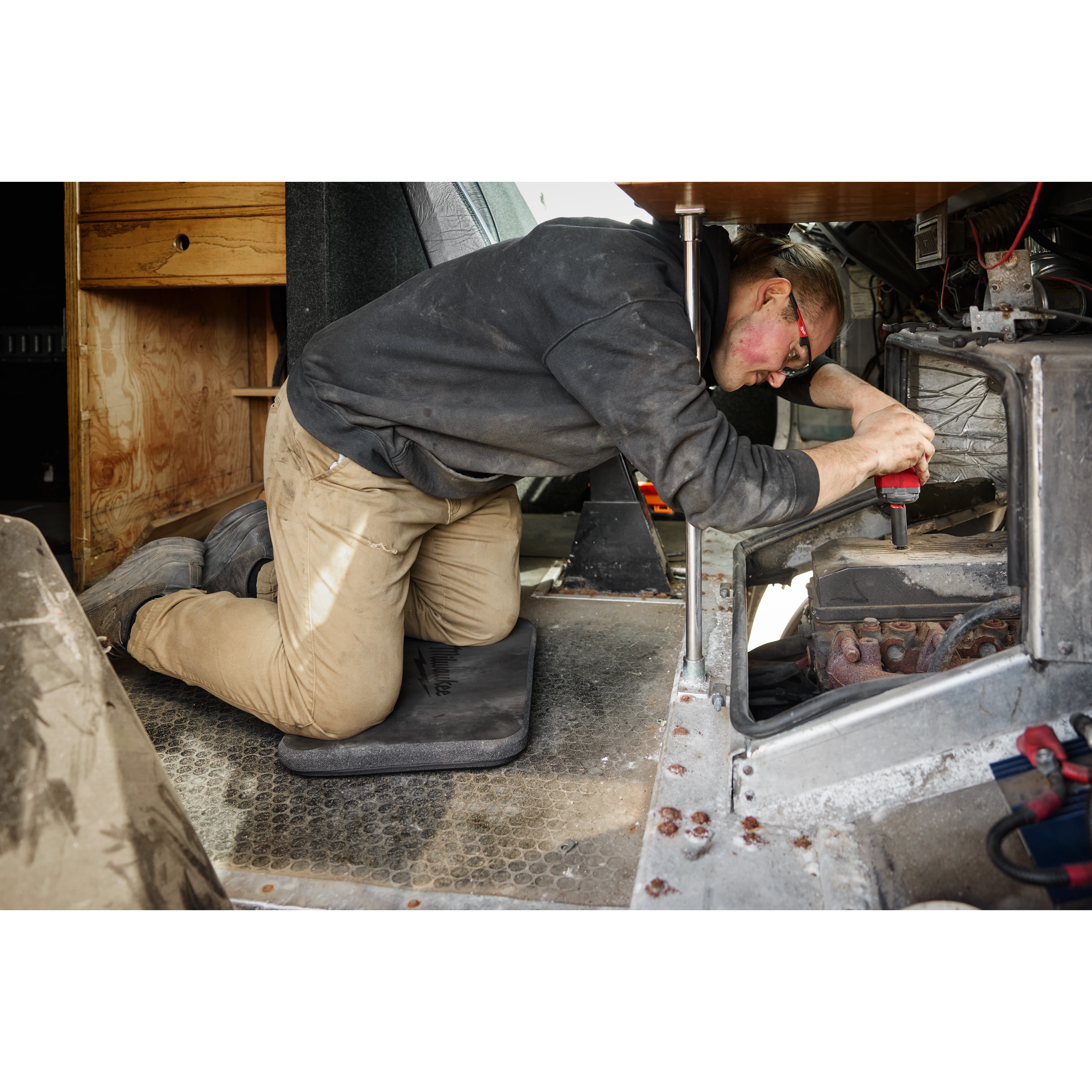 A person is working on a mechanical task, kneeling on a black Kneeling Pad. The surroundings appear industrial with various tools and equipment visible.