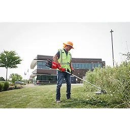 A person in a safety vest and gloves trims grass with a weed cutter outside a modern building.