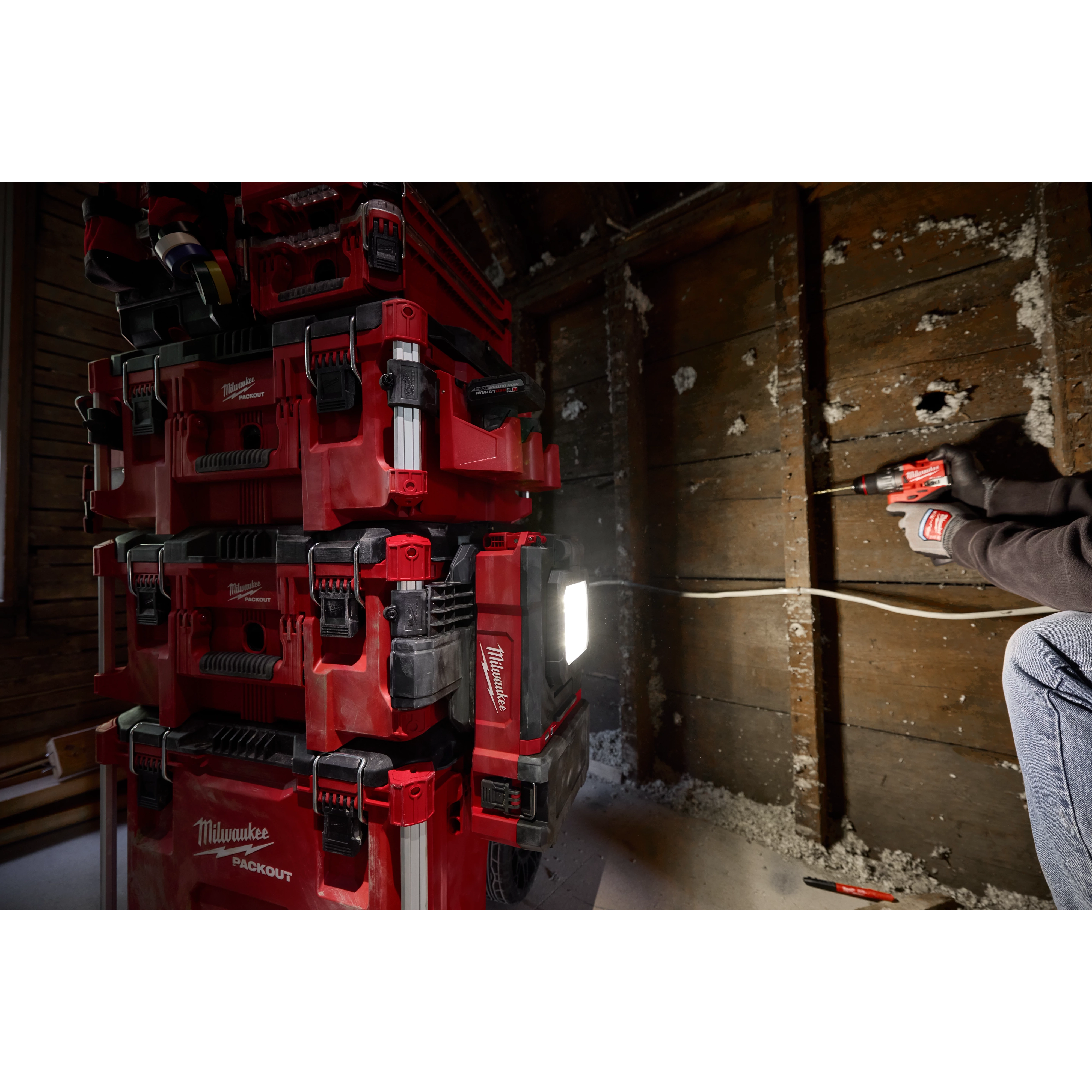 A stack of red Milwaukee PACKOUT™ Tool Boxes, including the PACKOUT™ Tool Box Compact Plate, is positioned against a wall with exposed studs. A person on the right uses a power drill on the wall, and a bright work light from the toolboxes illuminates the scene.