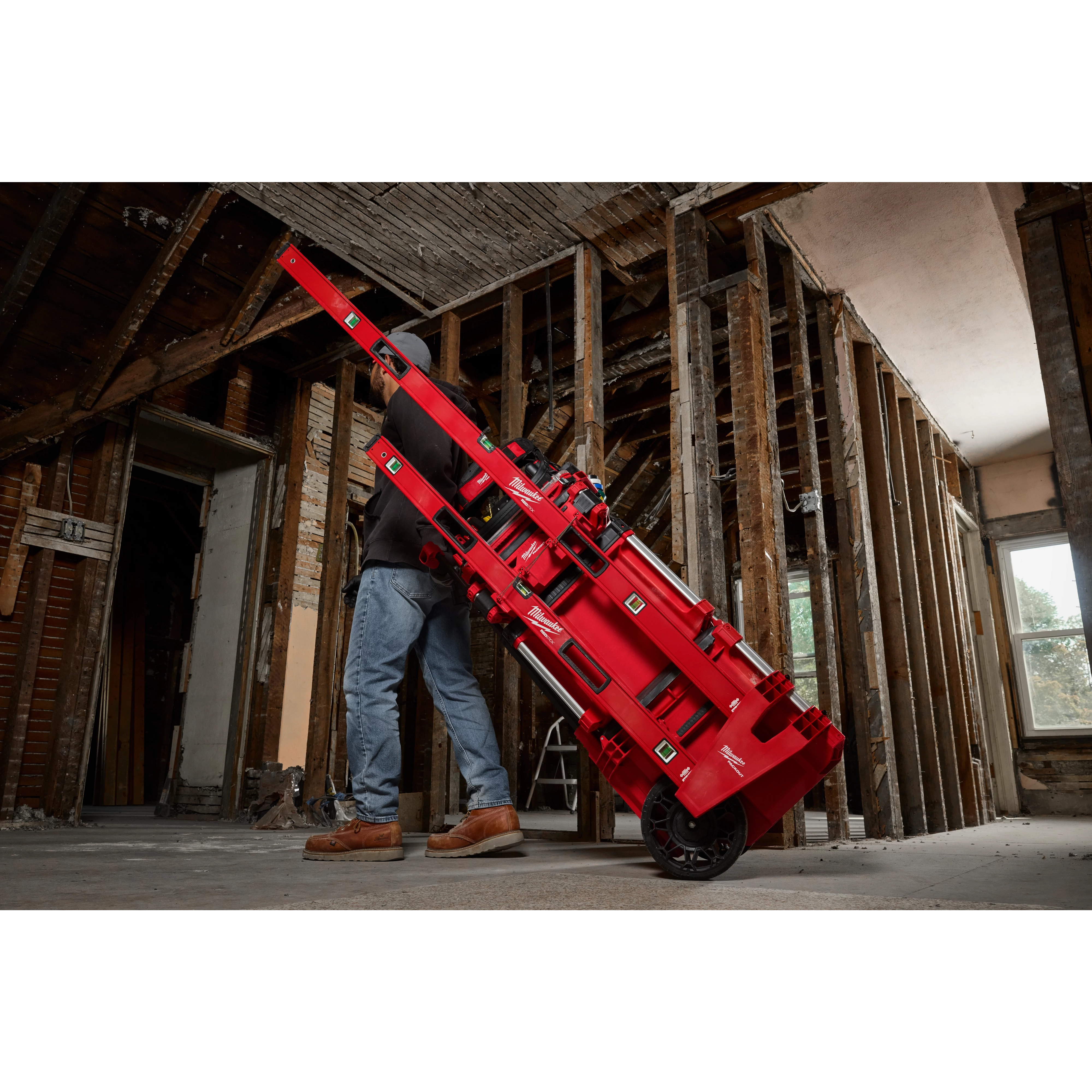 A person is pushing a red PACKOUT™ Tool Box with a Long Tool Holder Attachment through a construction site with exposed wooden beams. The tool box is stacked with other PACKOUT™ modules on a rolling cart.