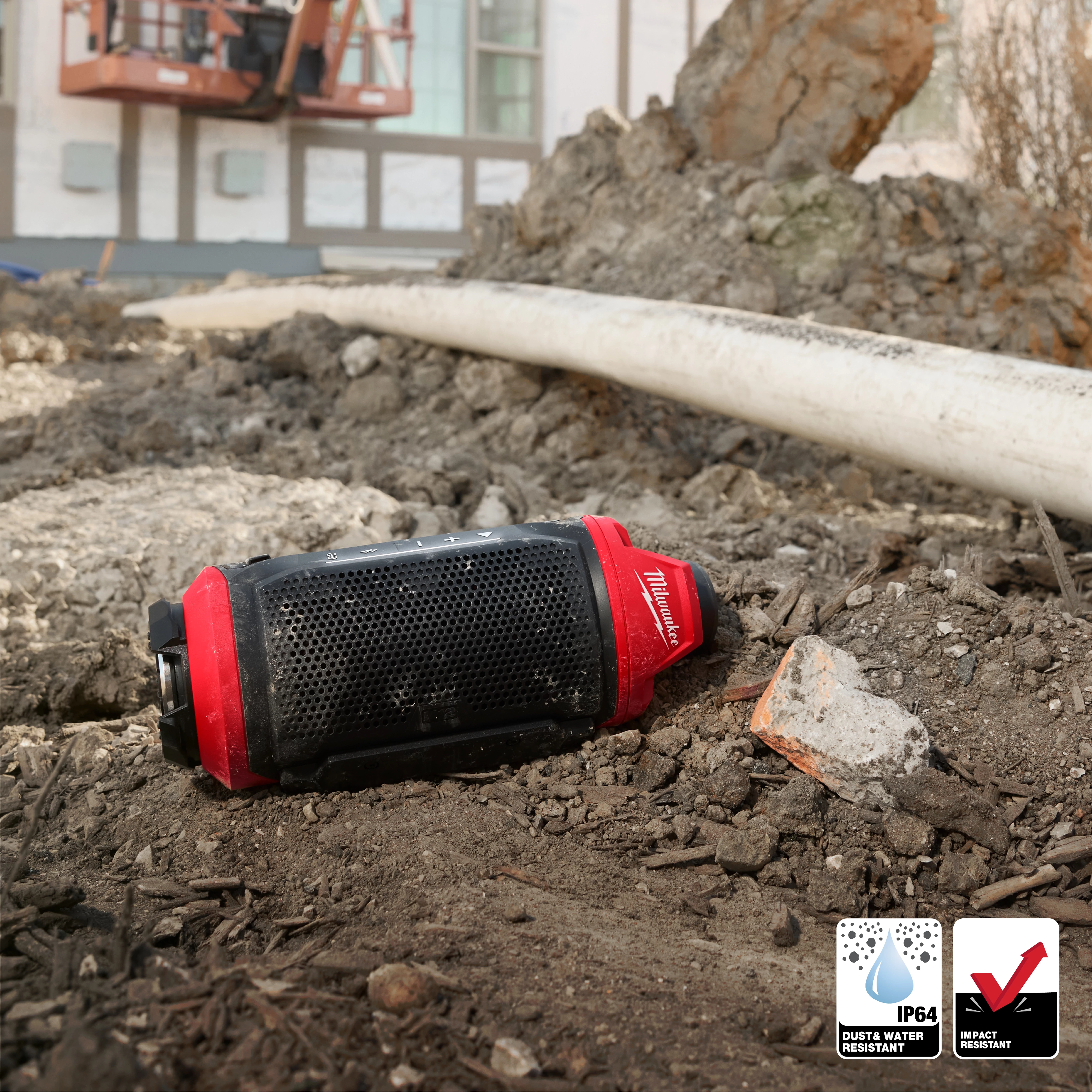 The image shows an M12™ Bluetooth® Jobsite Speaker w/ PACKOUT™ Compatibility lying on a rugged, outdoor construction site. The speaker is black with red accents and is positioned among dirt, rocks, and a pipe. Icons indicate its IP64 dust and water resistance, as well as impact resistance.