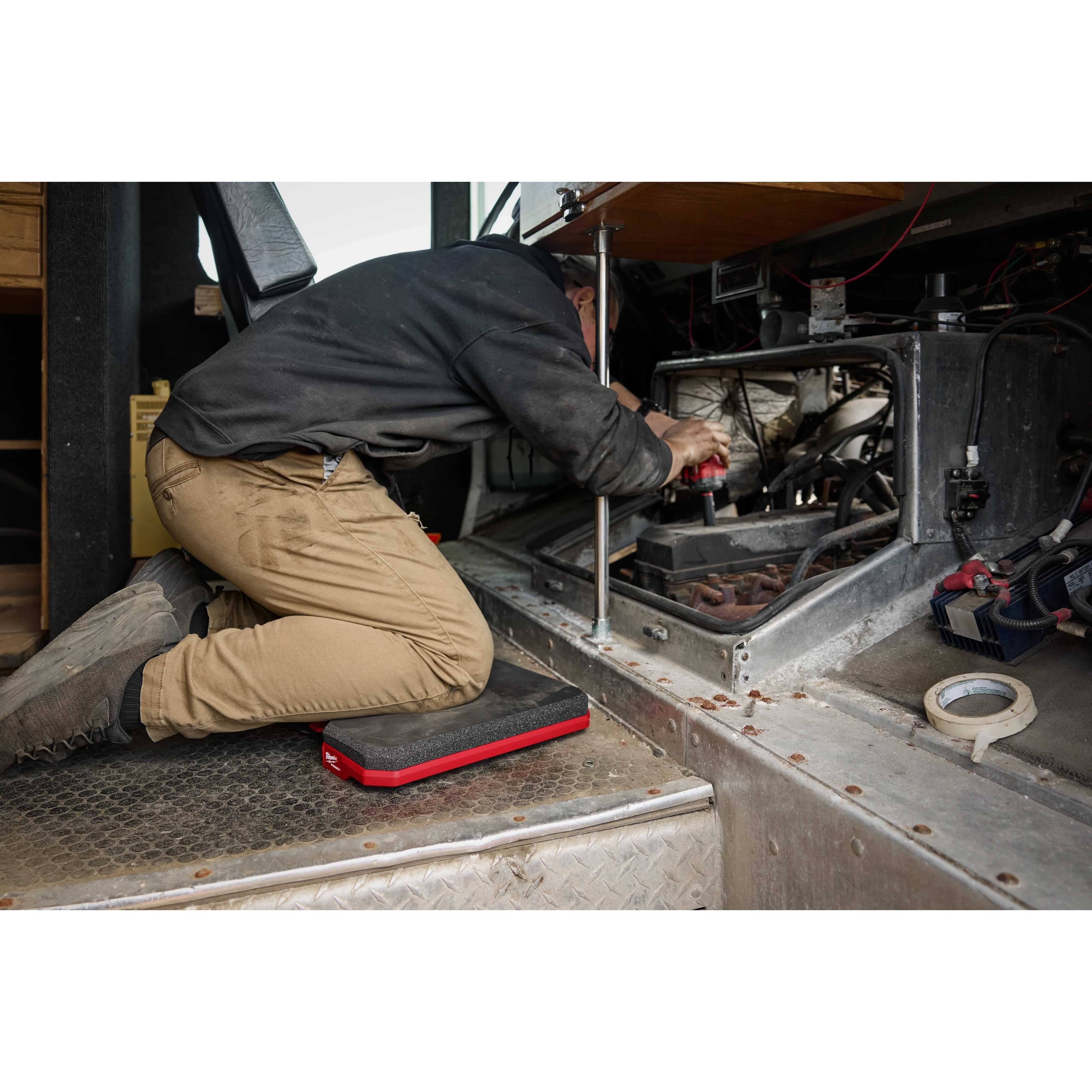 A person is kneeling on a PACKOUT™ Kneeling Pad while working on machinery inside a vehicle. The black and red knee pad provides cushion and support on the metal surface. Surrounding tools and equipment are visible in the background.