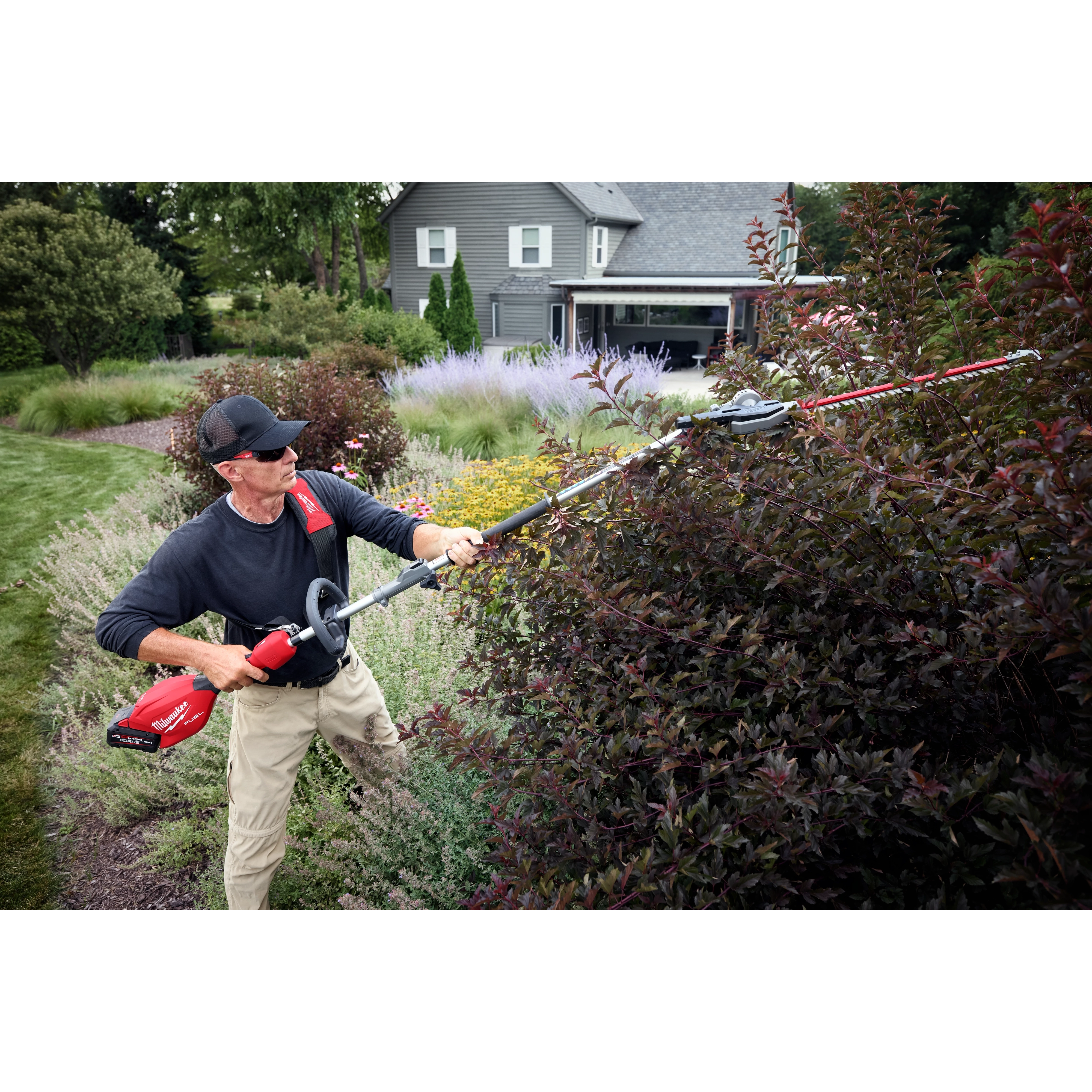 Person trimming bushes in a garden with a long-handled electric trimmer. House and various plants in the background.