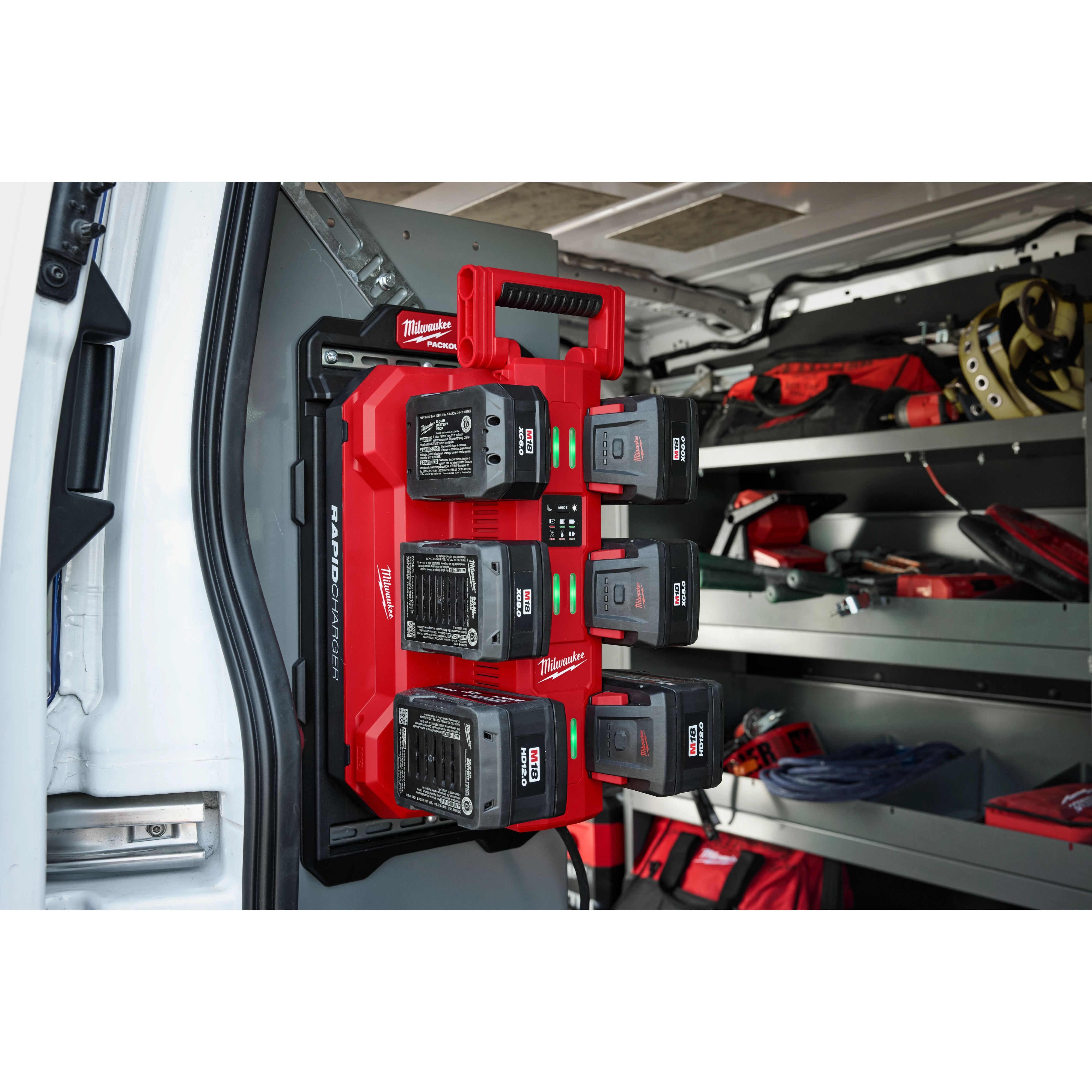 The image shows an M18™ Six Bay Daisy Chain Rapid Charger mounted inside a vehicle. The charger, red in color, is holding six batteries and is secured to the wall. Shelving units and various tools are visible in the background inside the vehicle.