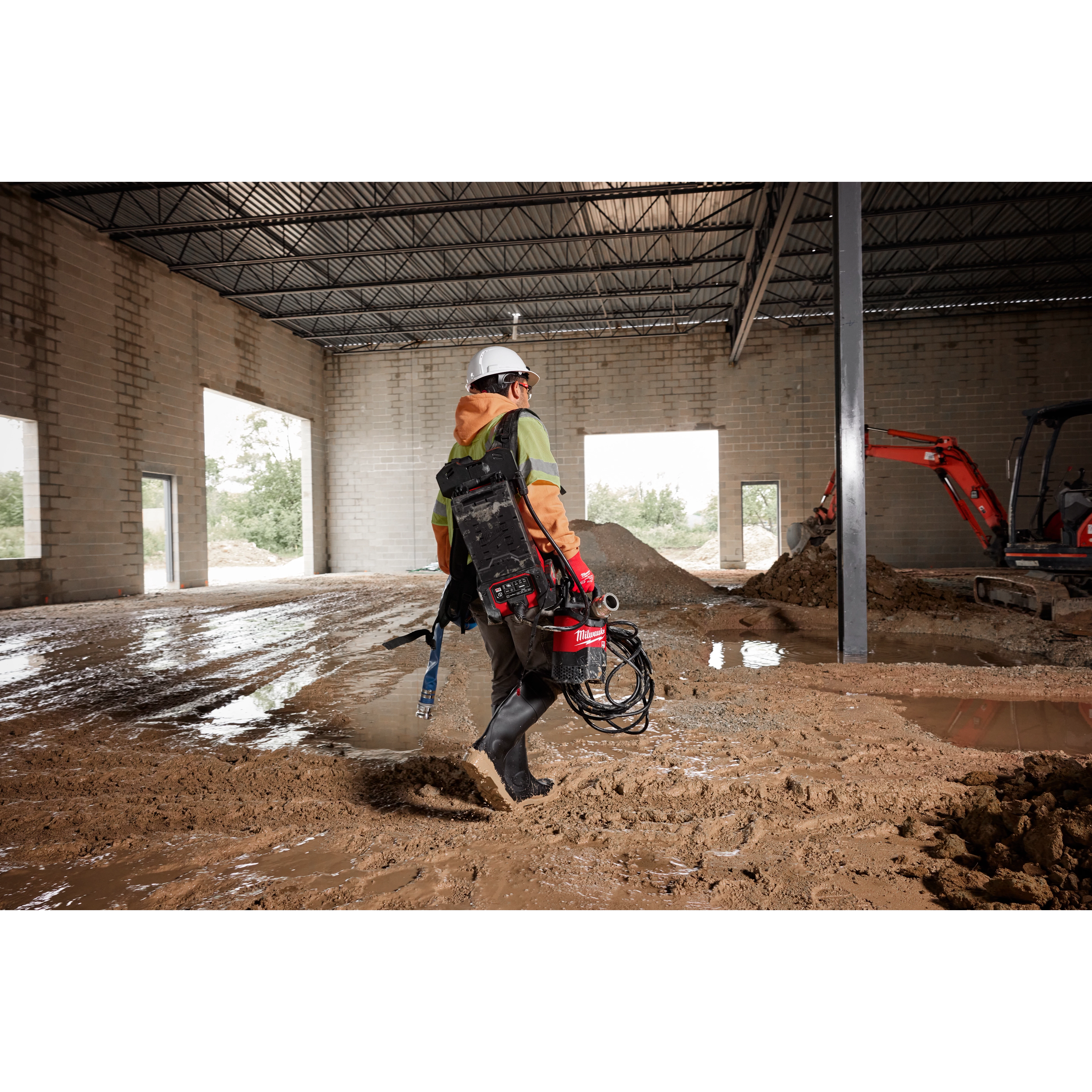 A person wearing a hard hat and protective gear carrying an MX FUEL™ Portable Pump Power Base and an MX FUEL™ 1HP 2" Submersible Pump Kit across a muddy construction site. The scene includes the interior of an unfinished building and construction equipment in the background.