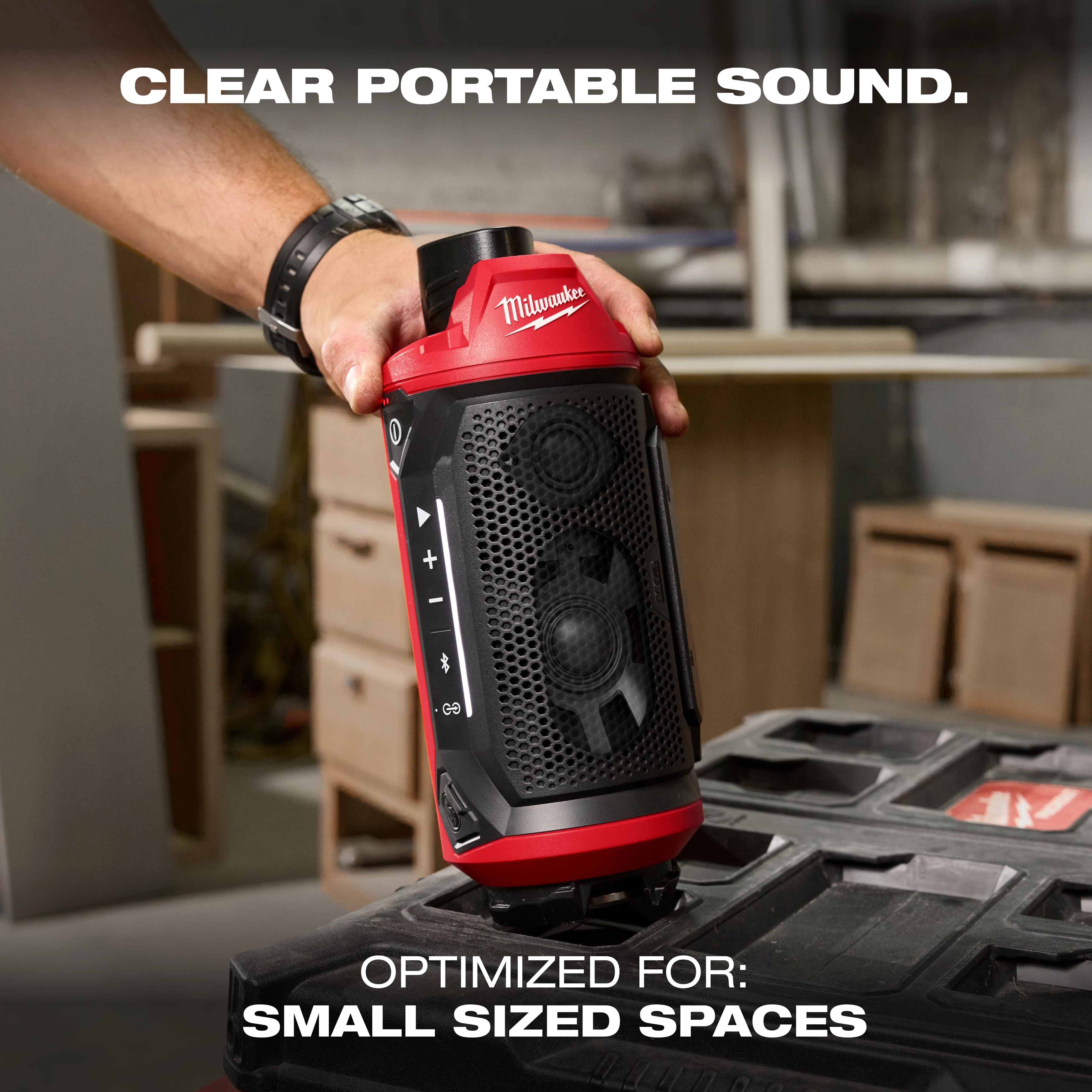 A person holds the M12™ Bluetooth® Jobsite Speaker w/ PACKOUT™ Compatibility in a workshop. The small, portable speaker is black and red, with control buttons on the side. The background includes shelves and tools, and the text "CLEAR PORTABLE SOUND" and "OPTIMIZED FOR: SMALL SIZED SPACES" is displayed.