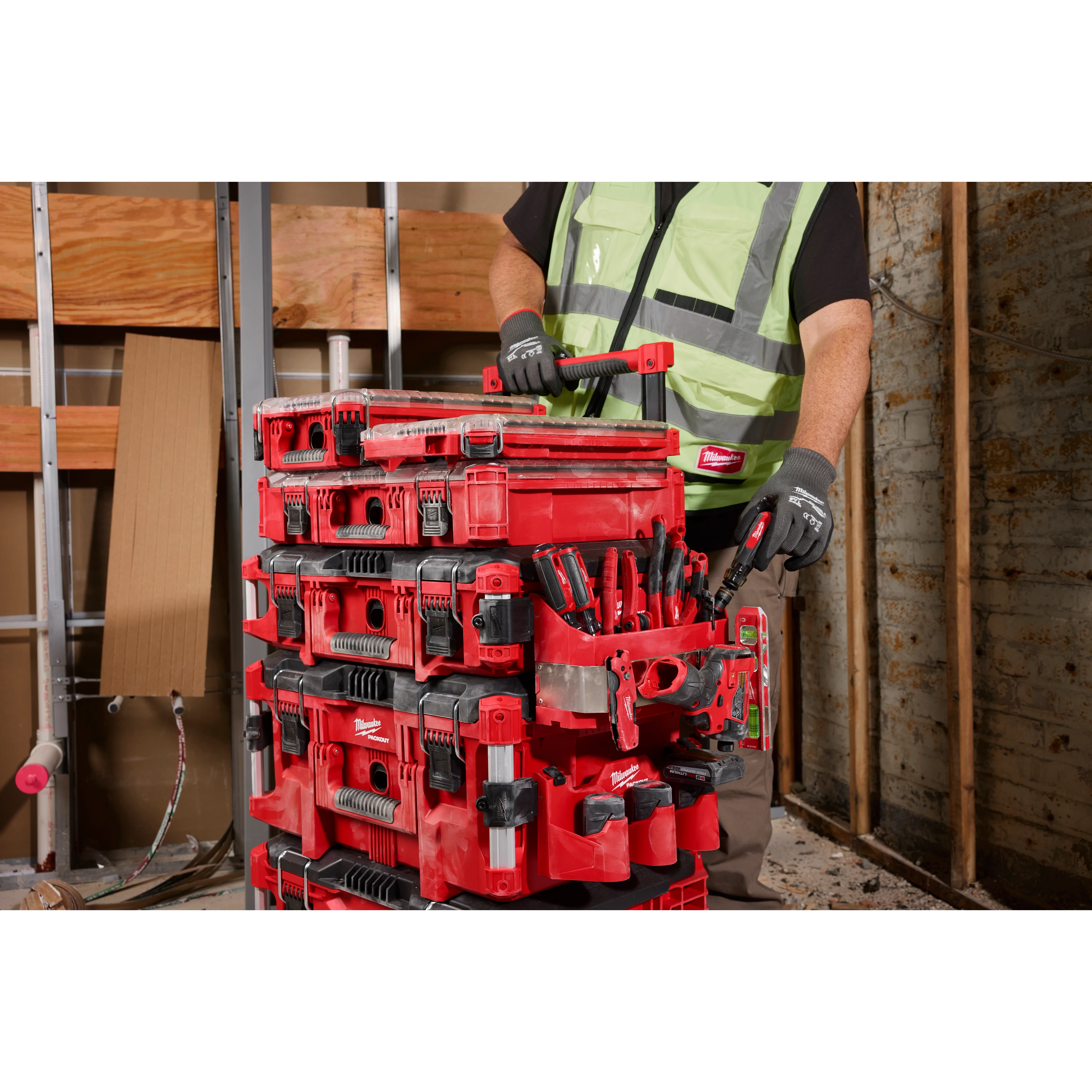 A person wearing work gloves and a high-visibility vest lifts a PACKOUT™ Tool Box Caddy Attachment in a workshop.