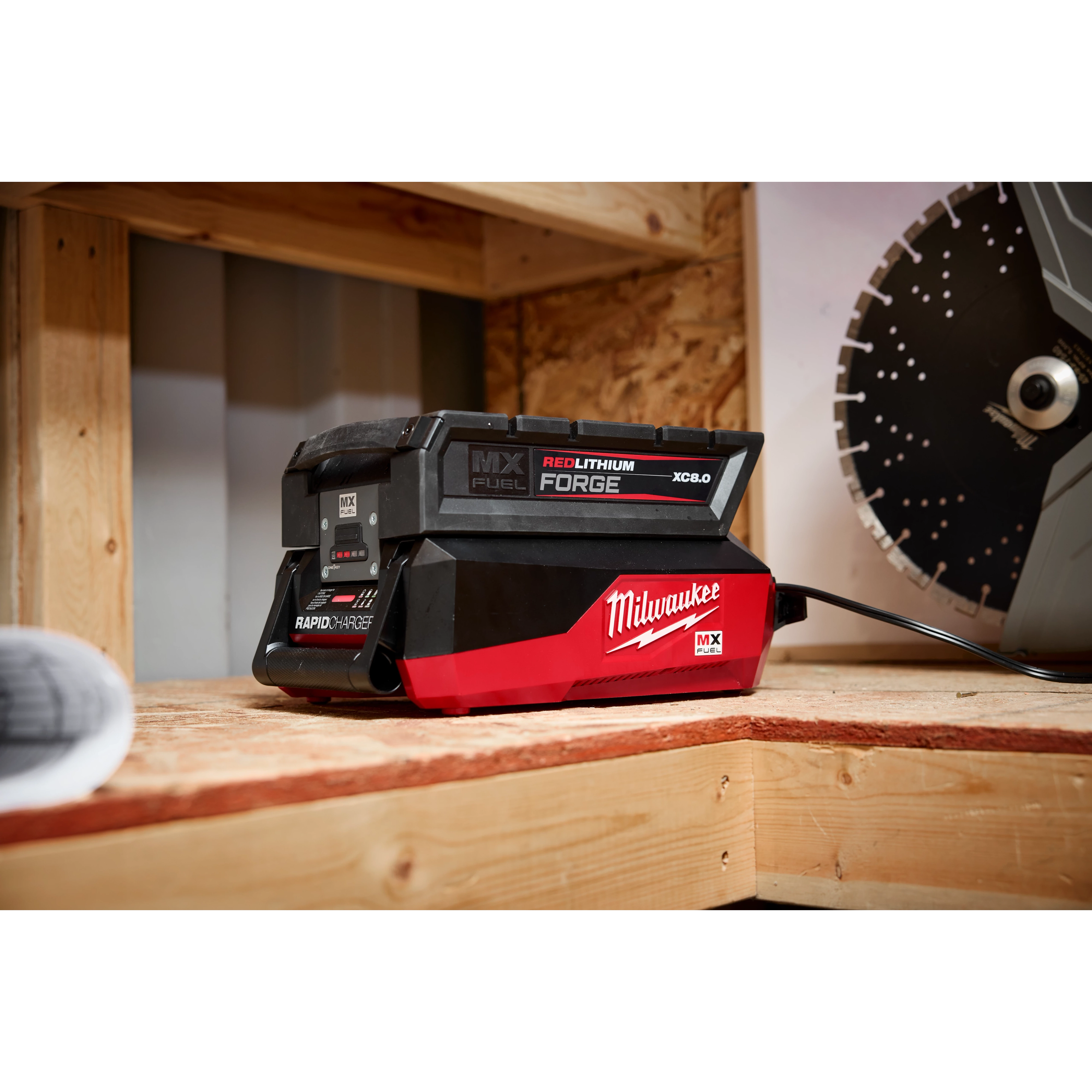 Milwaukee MX Fuel Redlithium battery charger on a wooden surface, with a charging indicator and a saw blade in the background.