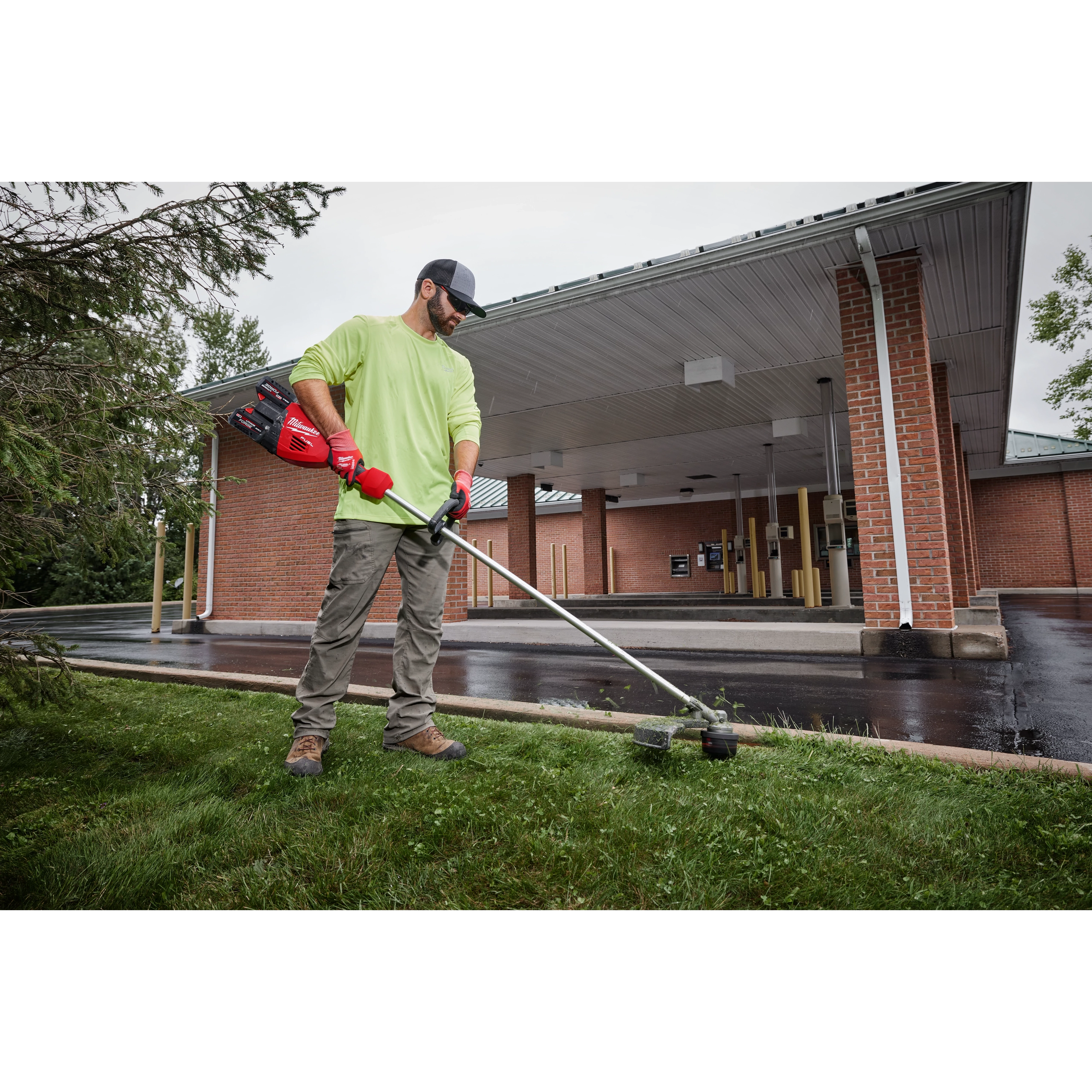 Person using a string trimmer to cut grass near a building with a covered driveway.