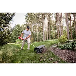 Person using a power tool to maintain a garden, surrounded by lush greenery and tall trees.