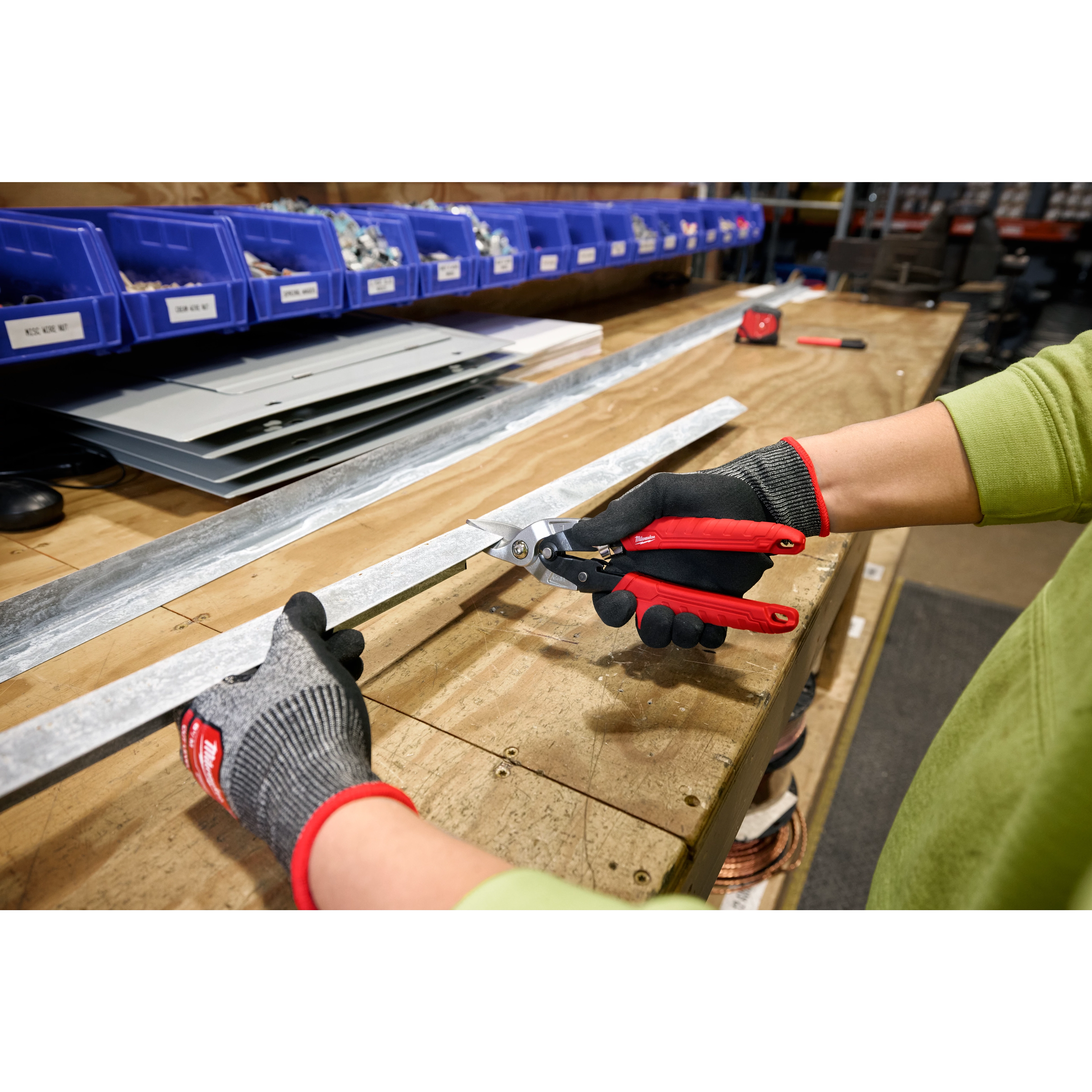 A person wearing gloves uses Left Cutting Aviation Snips to cut a piece of metal on a workbench. The workspace has blue bins with materials and tools, and there are metal sheets and other supplies nearby.