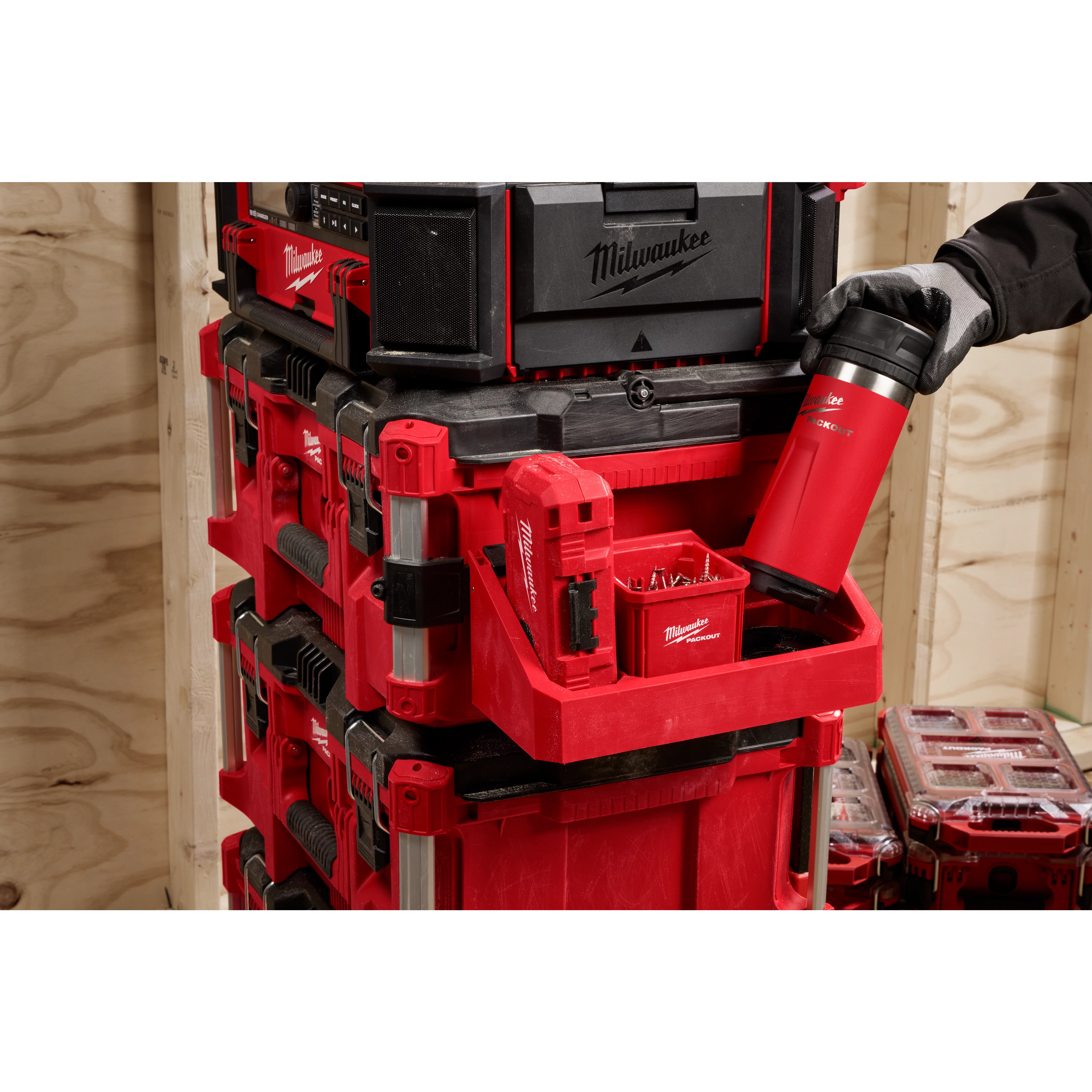 Stacked Milwaukee toolboxes with a hand placing a red thermos into a compartment, in a wooden workshop setting.