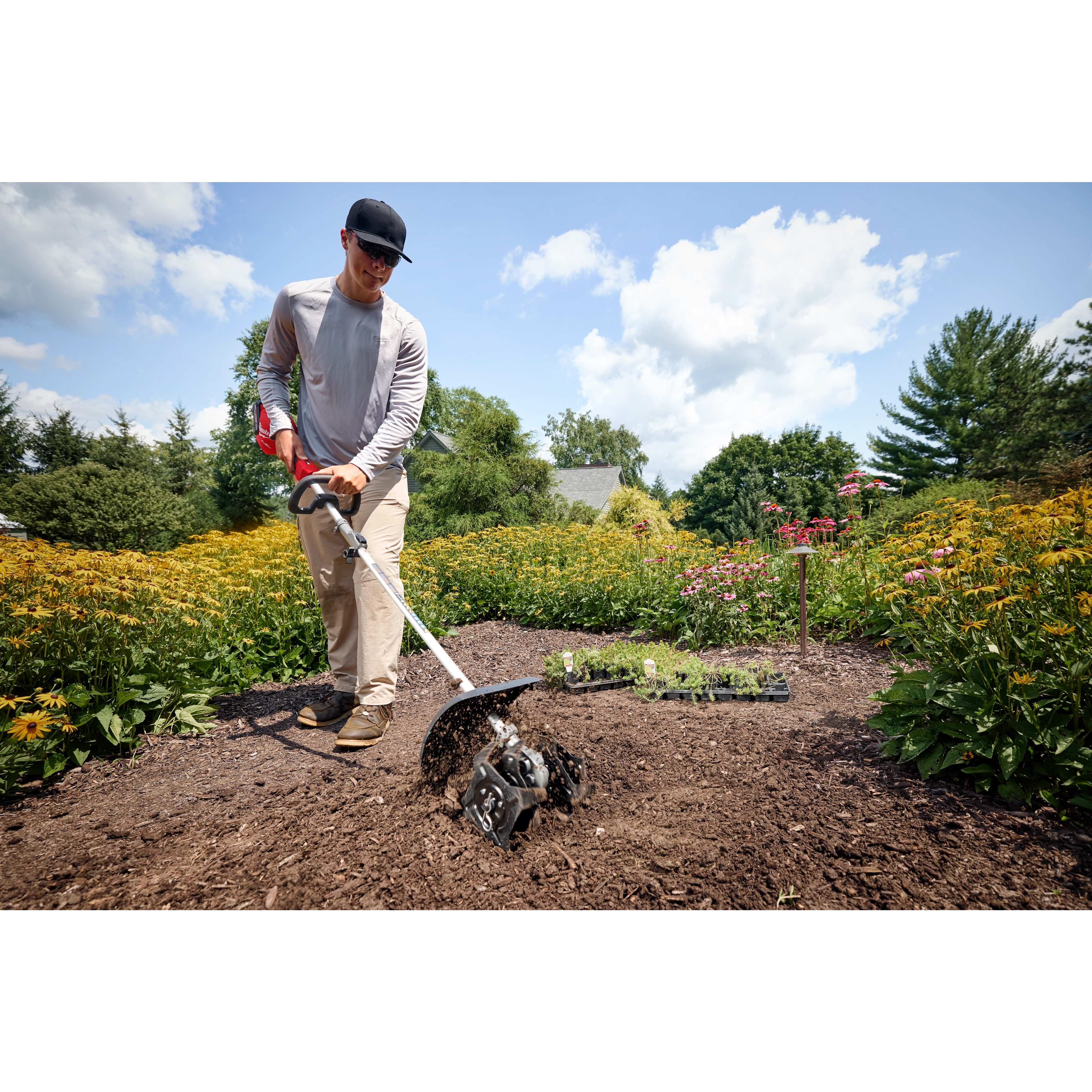 Person using a soil tiller in a garden surrounded by blooming flowers and bright green foliage.