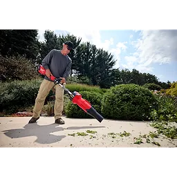 Person using a red leaf blower on a patio, surrounded by greenery and trees.