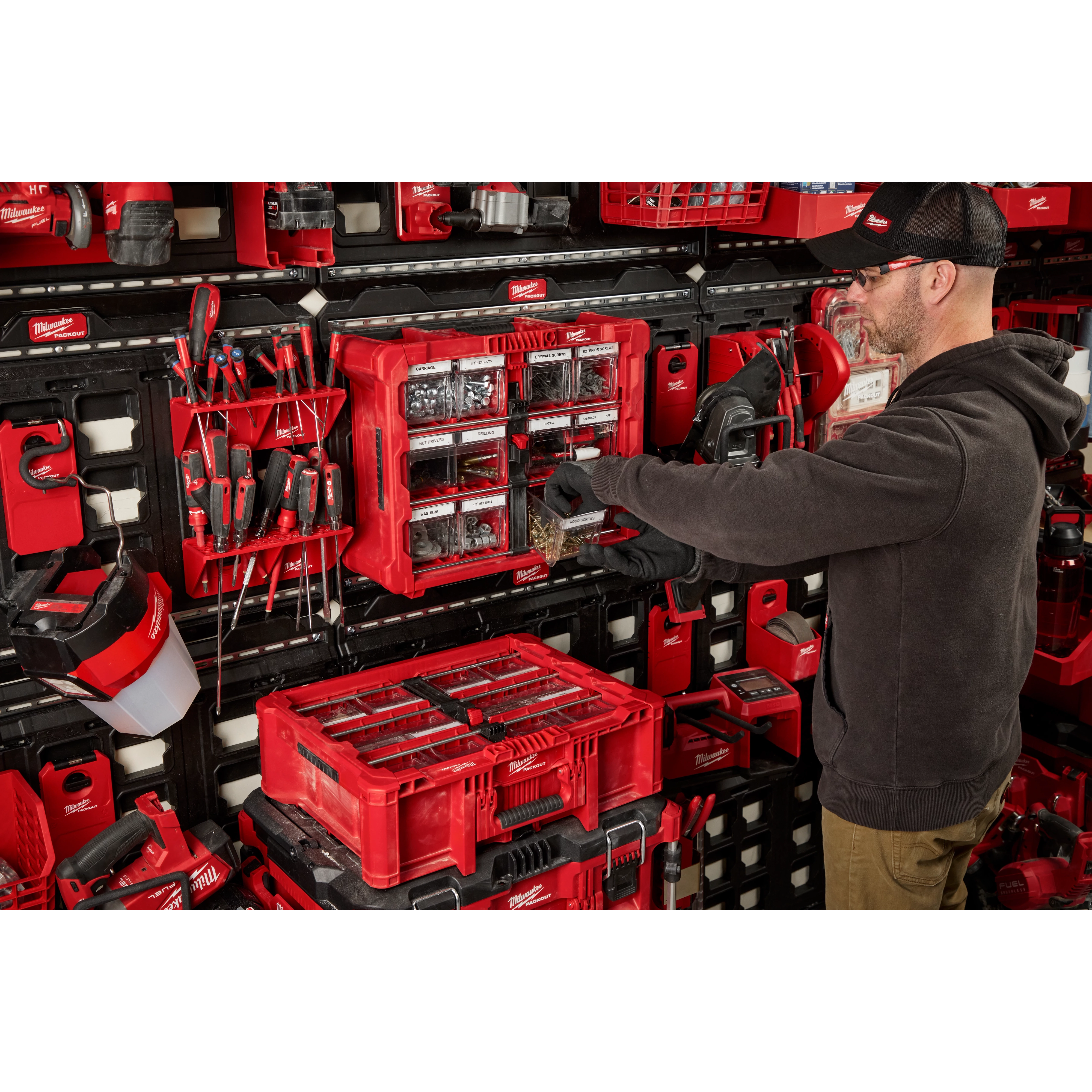 A person is organizing tools using the PACKOUT™ Tilt Bin Organizer. The organizer is mounted on a wall among various other PACKOUT™ storage products. It contains several small compartments filled with assorted items. The surrounding area includes more red storage units and various hand tools.