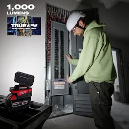 Person wearing a hard hat working with an electrical panel, illuminated by a Milwaukee TRUEVIEW light displaying "1,000 lumens."